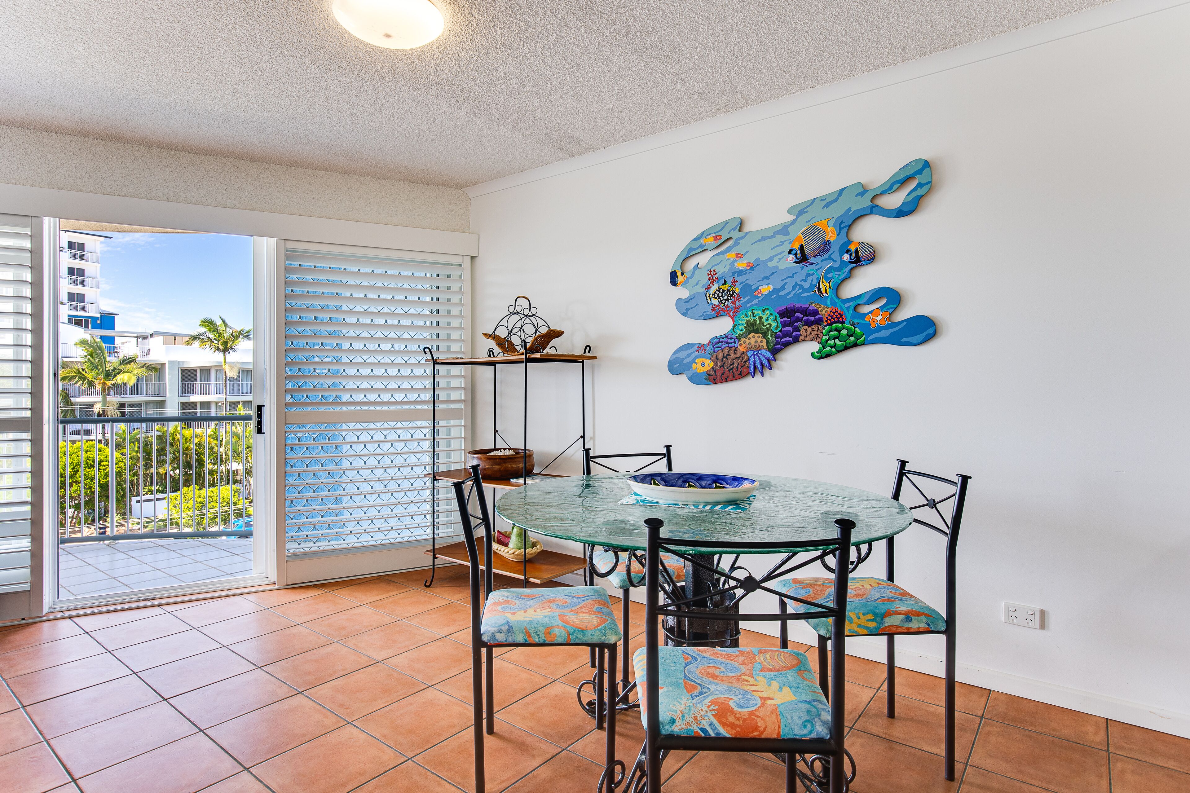 A dining area with a glass table and chairs, a decorative wall piece, and a view of a residential area through a sliding door.