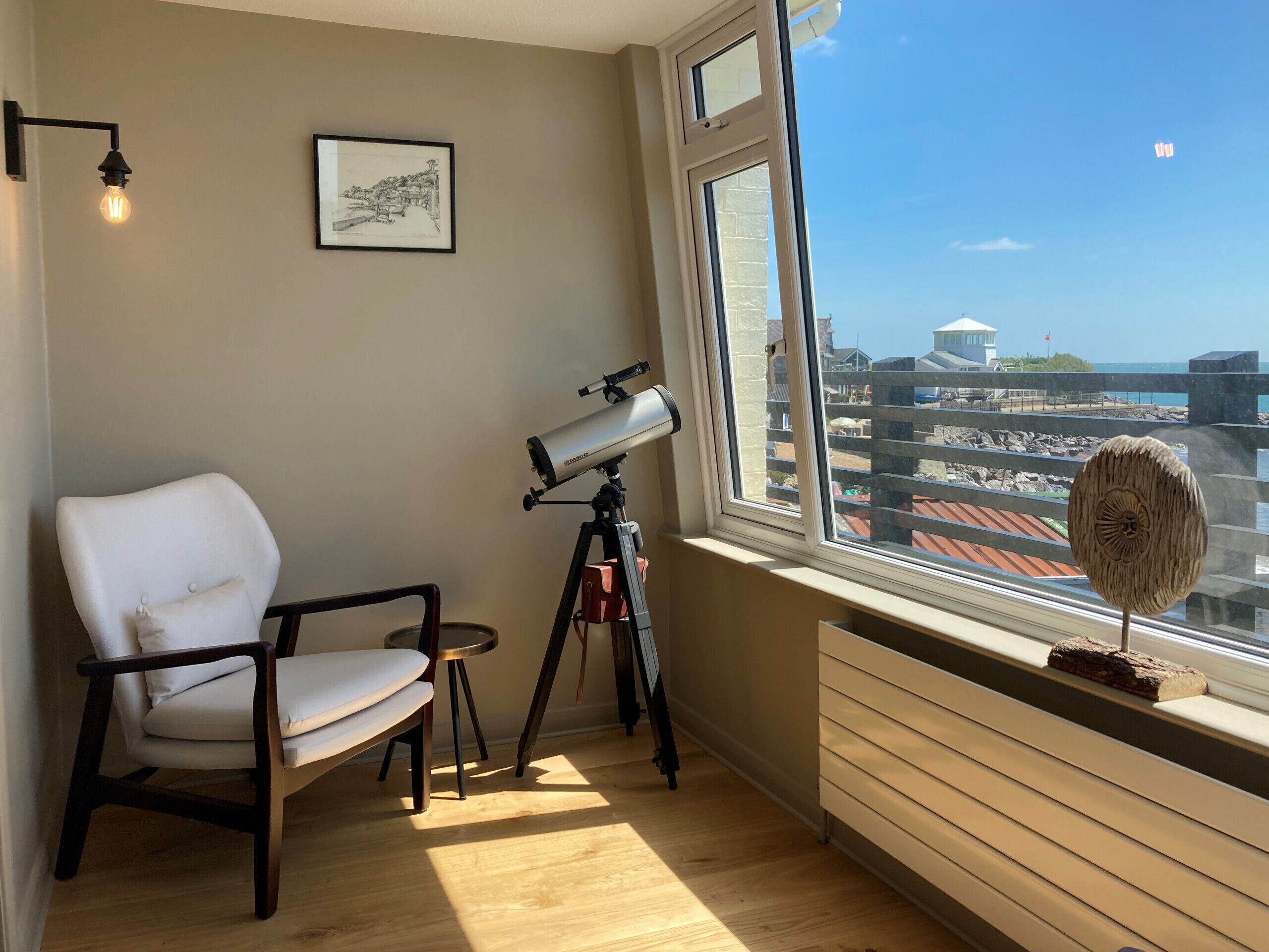 A room with a telescope on a tripod, a white chair, a framed picture on the wall, and a view of buildings through a large window.