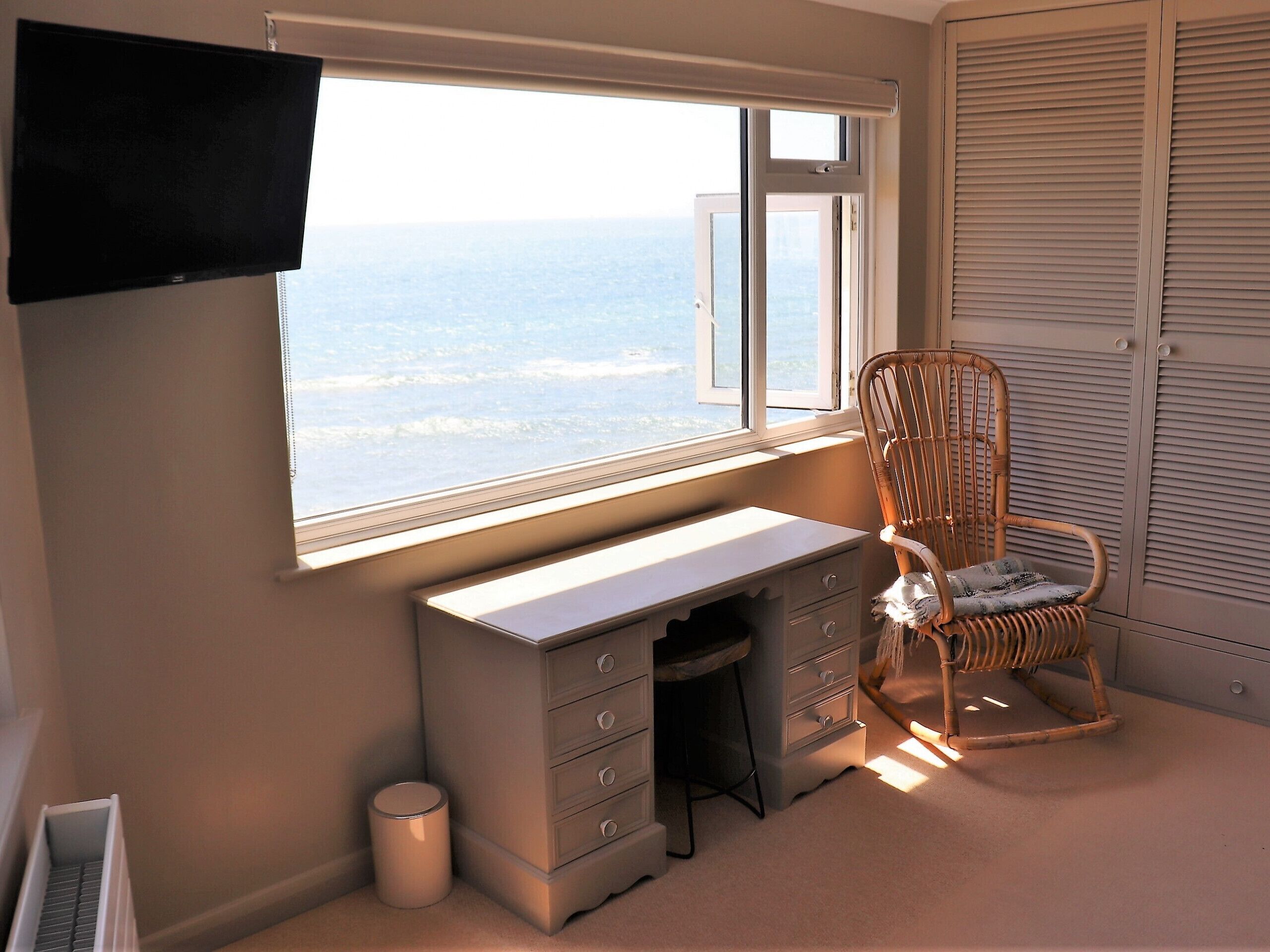A bedroom with a desk, a chair, a window with a view of the sea, and a wardrobe.