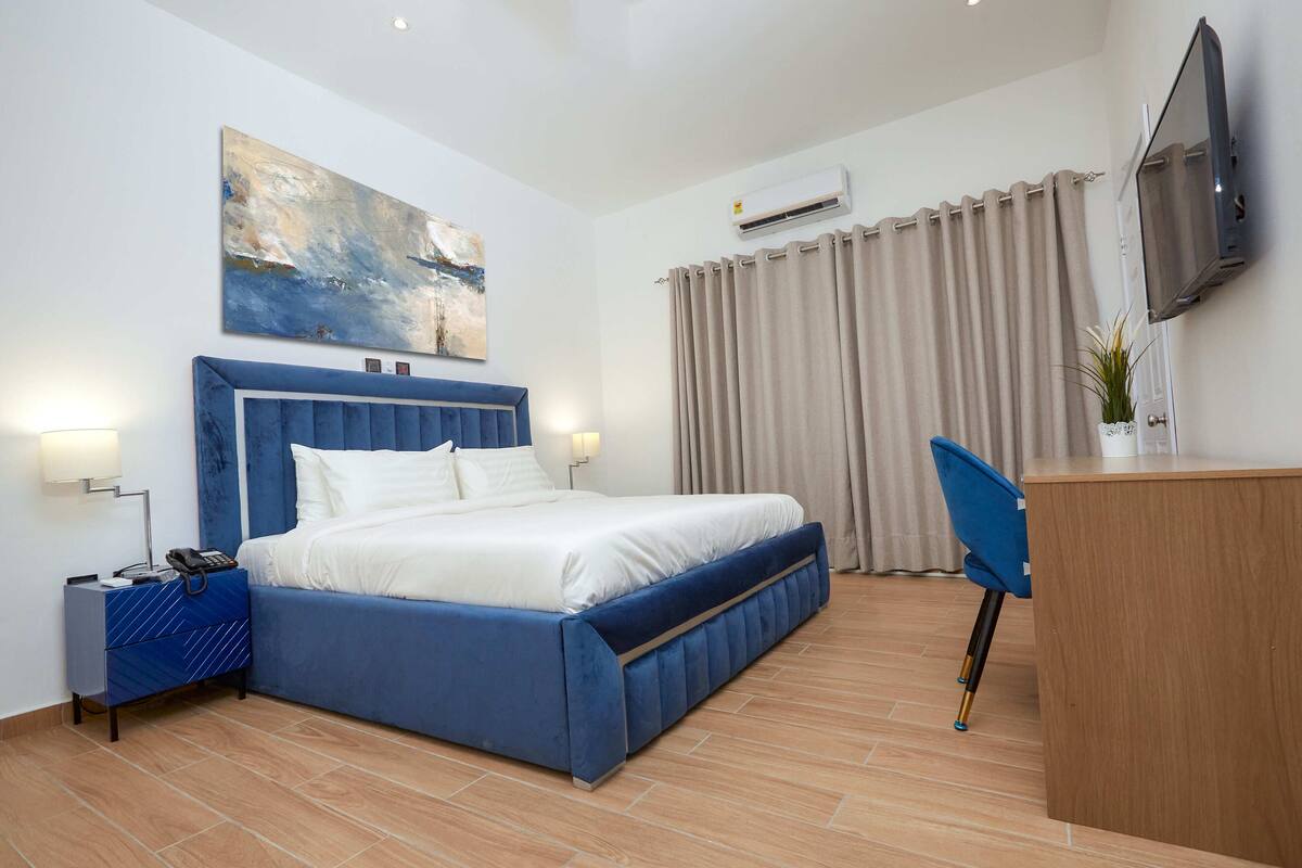 A hotel room with a large bed, a desk, a chair, a TV, and an air conditioning unit.