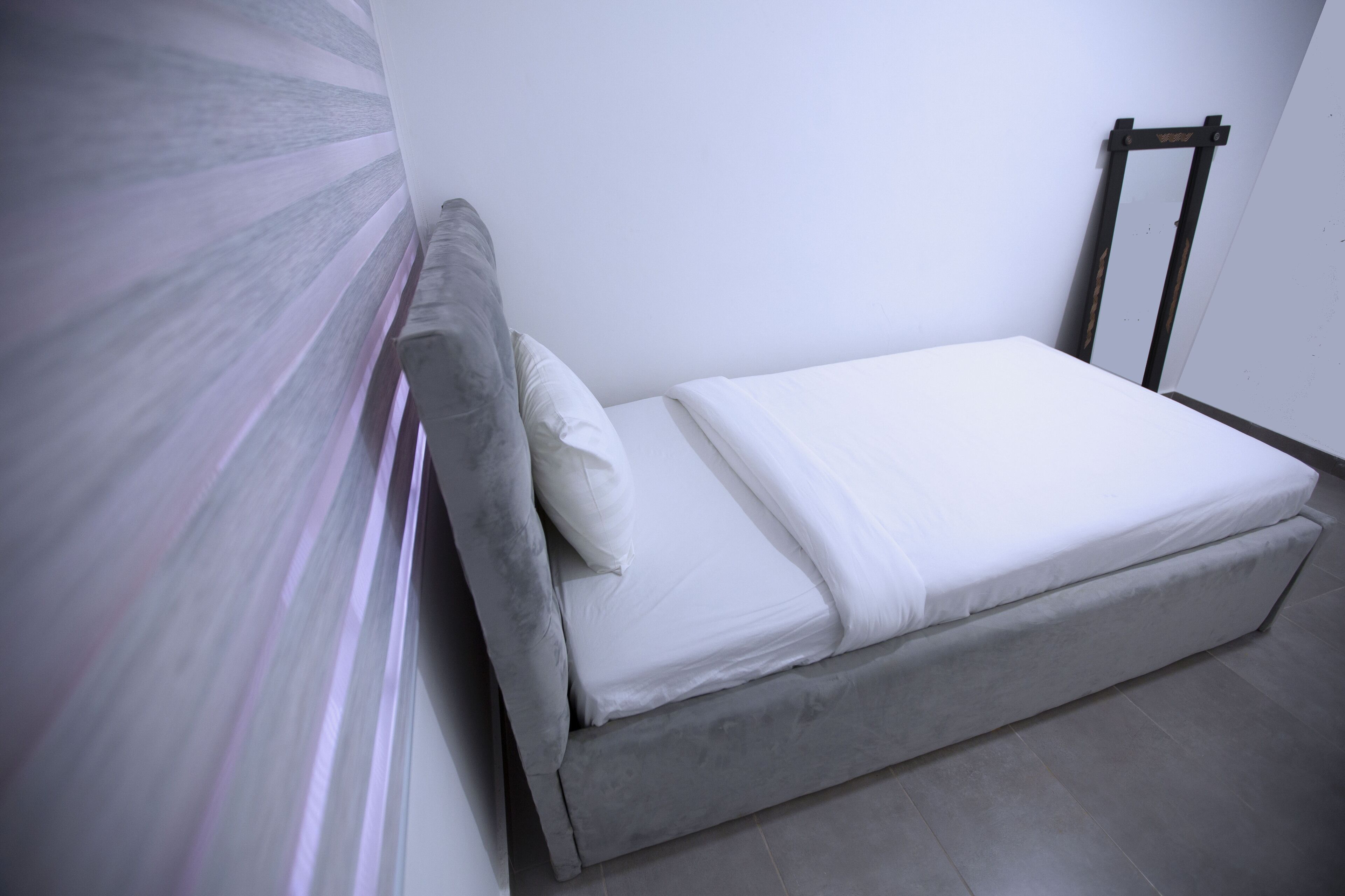 Una cama individual con cabecera gris y ropa de cama blanca en una habitación con paredes blancas y suelo gris.