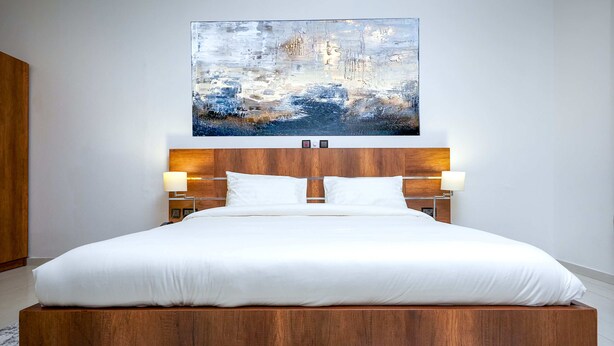A modern bedroom with a wooden headboard, white bedding, and a large abstract painting above the bed. - Maple Homes (Accra)