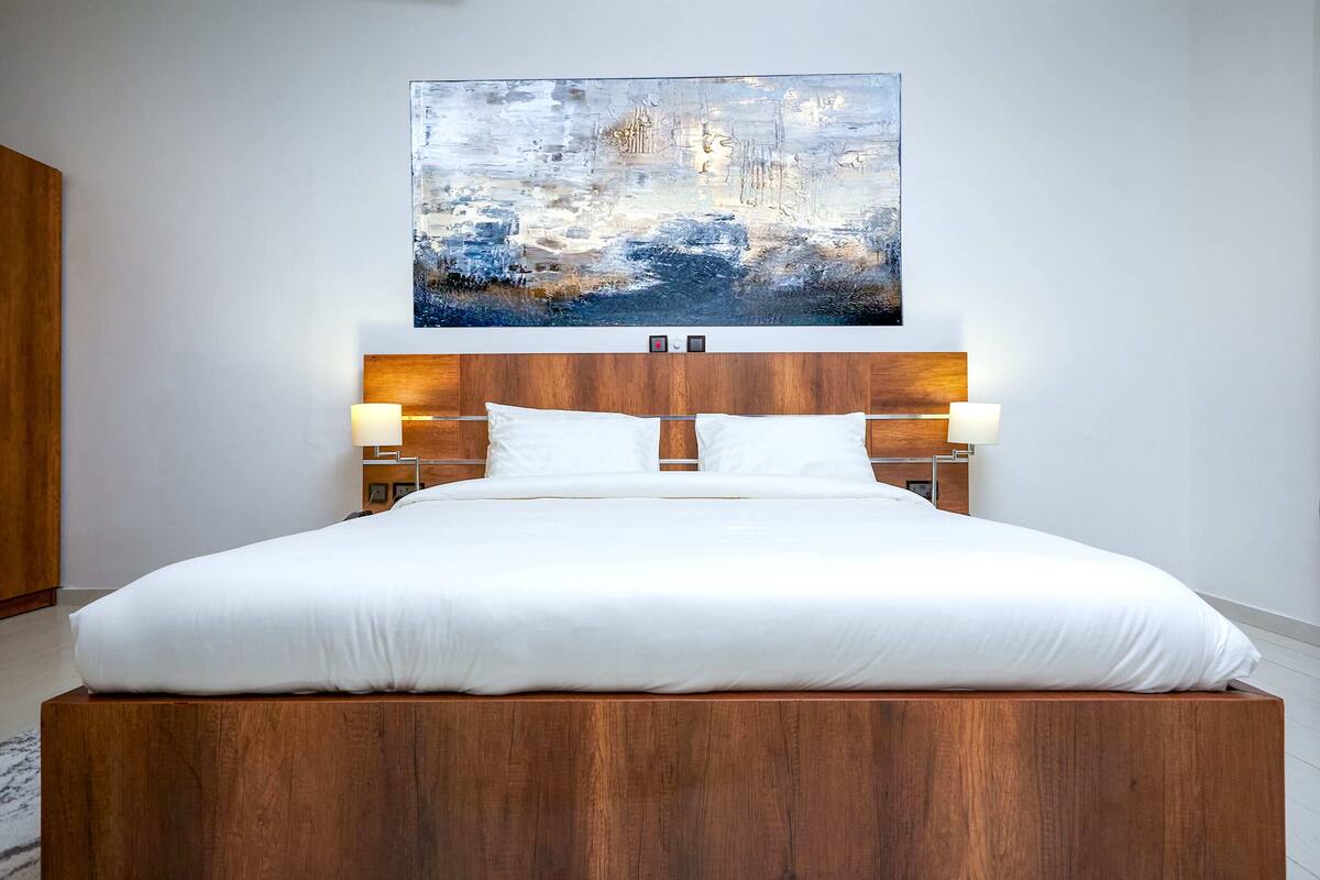 A modern bedroom with a wooden headboard, white bedding, and a large abstract painting above the bed.