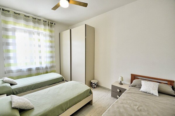 A bedroom with two single beds, a bedside table with a lamp, a small trash bin, and a ceiling fan.