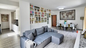 A modern living room with a grey sectional sofa, a large bookshelf, and a dining area with a table and chairs.