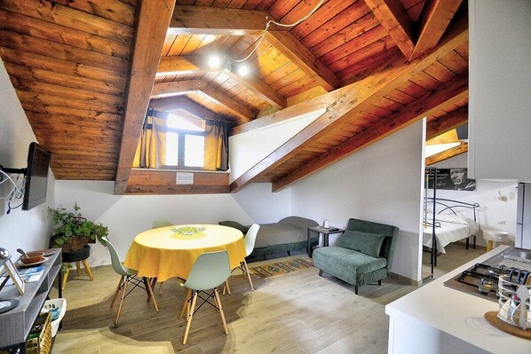 A cozy loft-style room with a high ceiling, wooden beams, a dining area with a yellow table, and a green sofa.