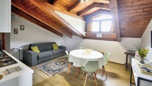 A cozy loft-style room with a high wooden ceiling, a dining area with a round table and chairs, a gray sofa with green cushions, and a kitchen area with a stove. - Villa Olga E: A cozy and welcoming apartment that is part of a villa situated in a quiet location, a few minutes from the town center, with Free WI-FI. (Agropoli)