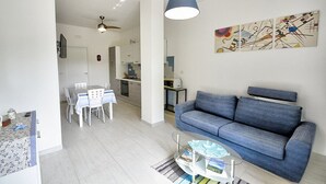 Un salon moderne avec un canapé, une table à manger et des chaises, un ventilateur de plafond, et une cuisine en arrière-plan.