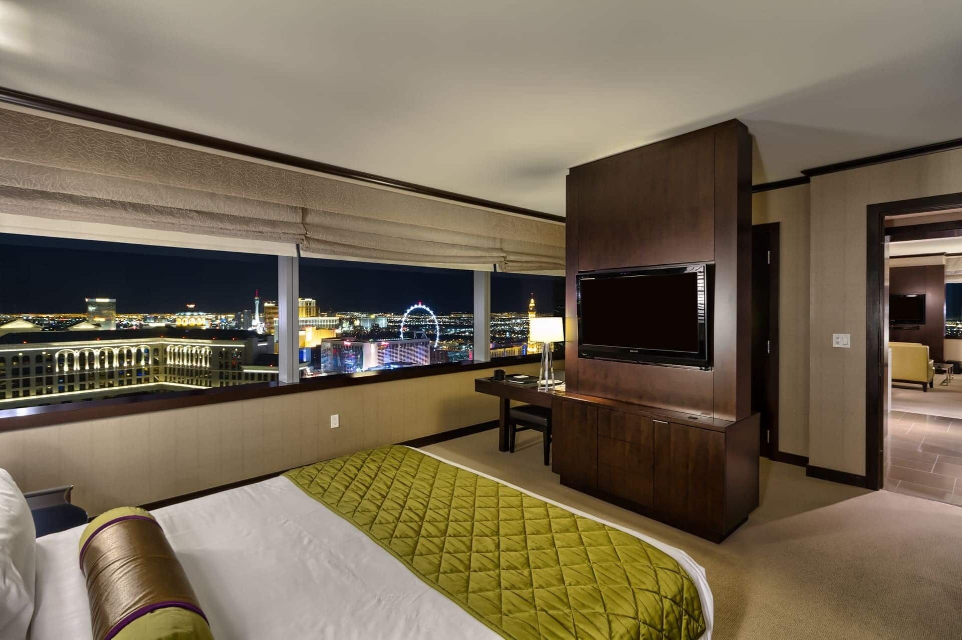 A hotel room with a large bed, a desk with a chair, a flat-screen TV mounted on the wall, and a view of the cityscape at night.