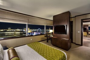 Habitación de hotel con una cama grande, un escritorio con silla, un televisor de pantalla plana en la pared y vista a la ciudad de noche.