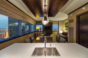 A modern hotel room with a large window offering a city view, a dining area with a white table and chairs, and a flat-screen TV mounted on the wall.