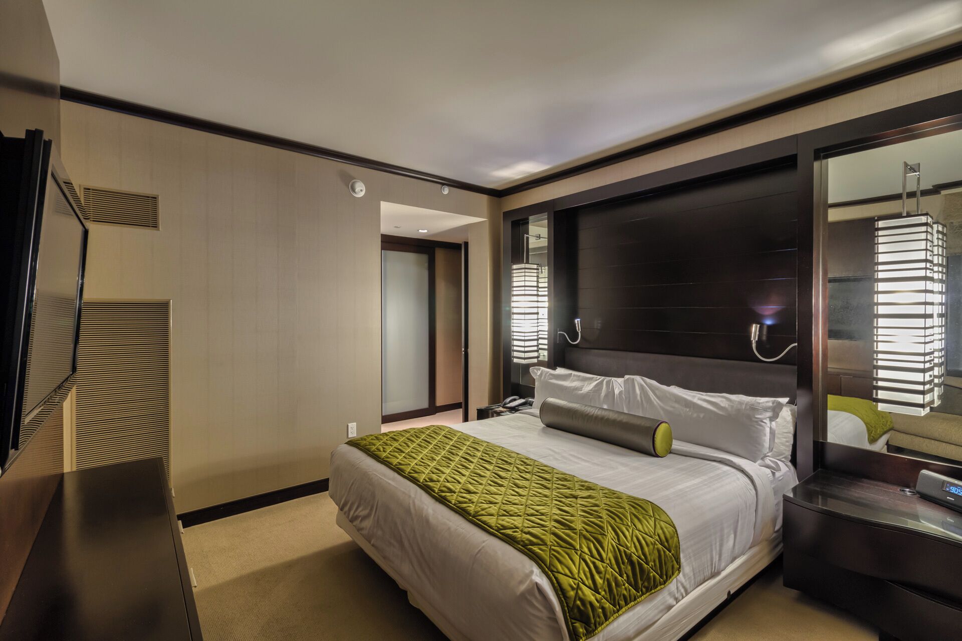 A modern hotel room with a large bed, a bedside table, a mirror, and a view of the hallway.