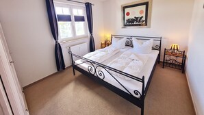 A bedroom with a metal bed frame, white bedding, a radiator, a window with curtains, a bedside table with a lamp, and a framed artwork on the wall.