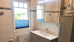 A bathroom with a window, white sink, mirror, and a refrigerator.