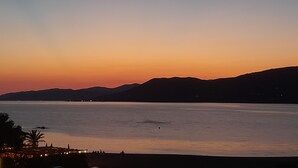 Vista de un atardecer costero con mar tranquilo, montañas lejanas y algunas personas en la orilla.