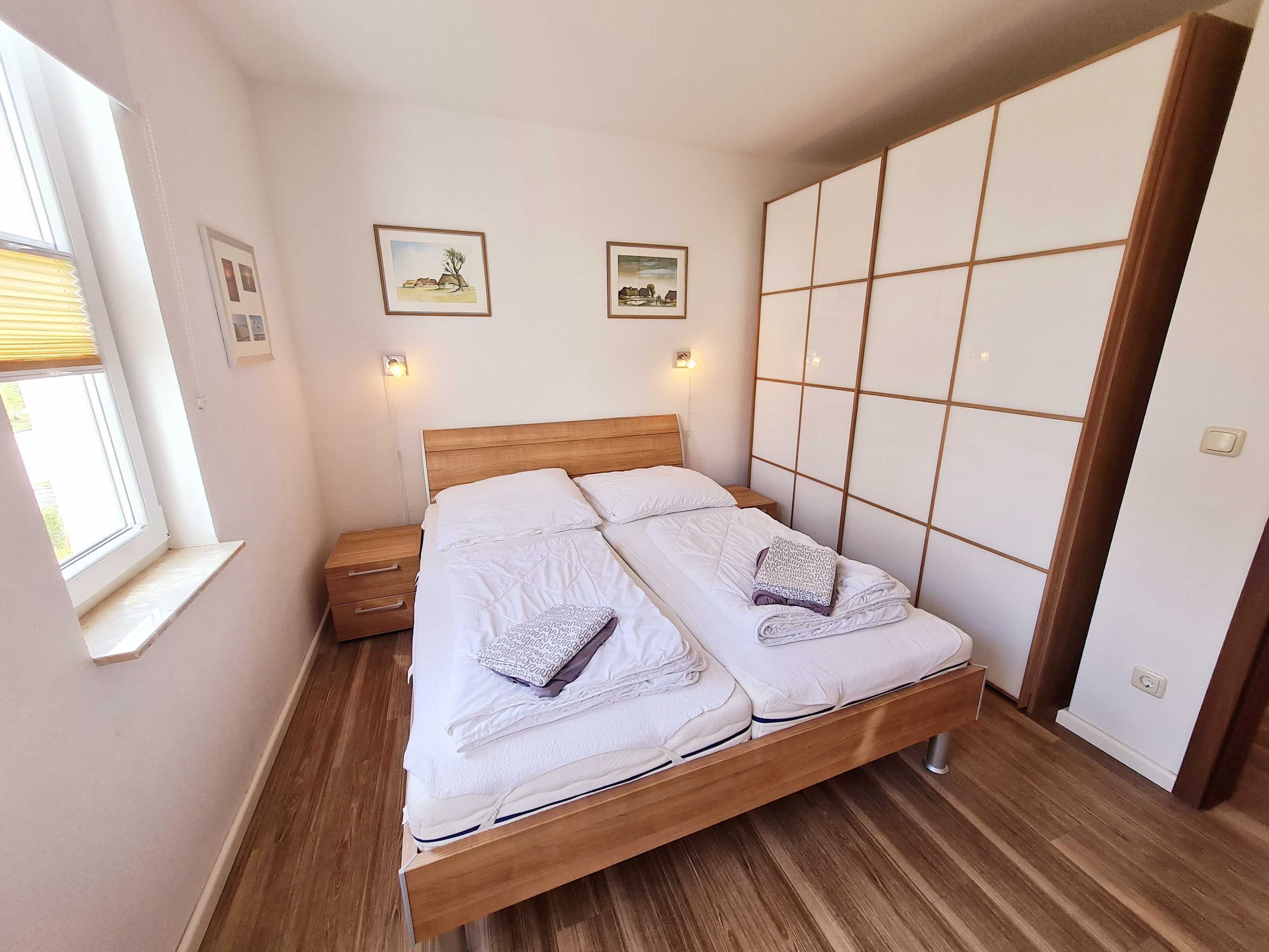 Ein Doppelbett mit Holz Kopfteil, zwei Nachttische, ein Fenster mit Jalousien und zwei gerahmte Bilder an der Wand.