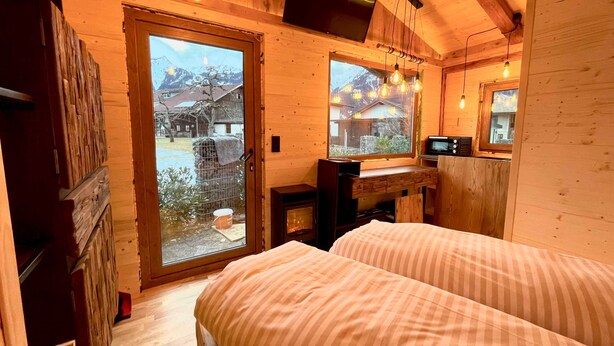 Habitación de cabaña de madera con cama, televisor, chimenea y ventana con vistas a una casa y montañas.