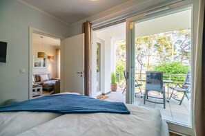 Une chambre d’hôtel avec un lit, un canapé, une table à manger et des chaises. Il y a un balcon avec vue sur les arbres et une chaise.