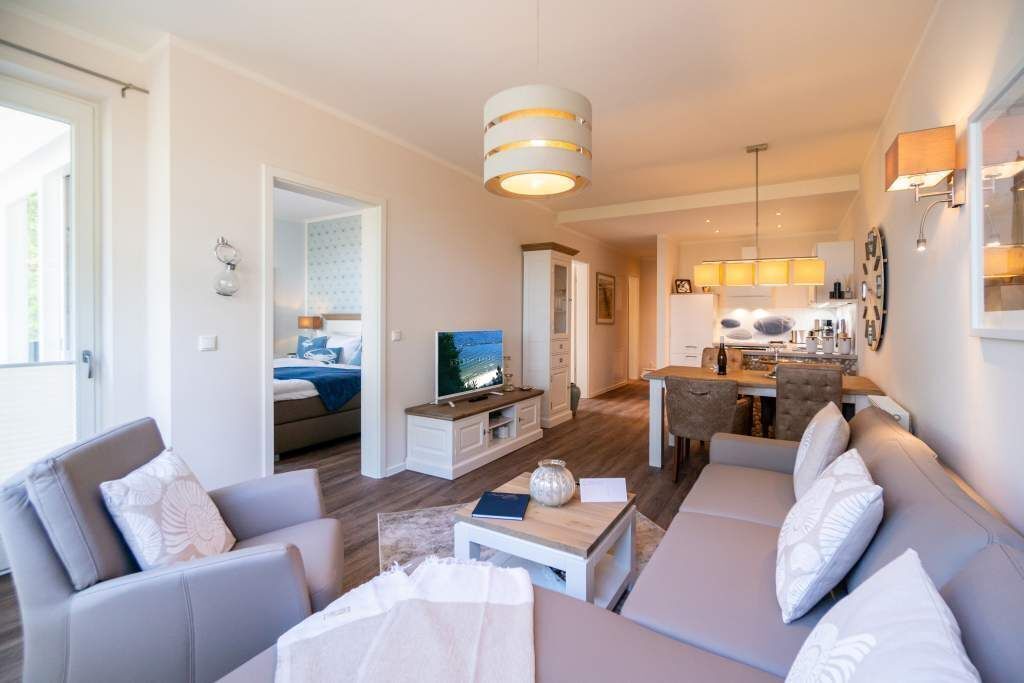 Una sala de estar moderna con sofá, mesa de centro y televisor. Hay un comedor con mesa y sillas. La habitación tiene una paleta de colores neutros y un ventanal grande.