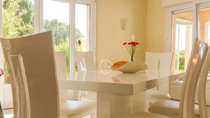 A dining area with a white table and chairs, a vase with flowers, and a view of greenery outside. - Peaceful Escape in Argeliers (Argeliers)