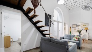 A modern living room with a grey sofa, wooden staircase, and a dining area with a white table and chairs.