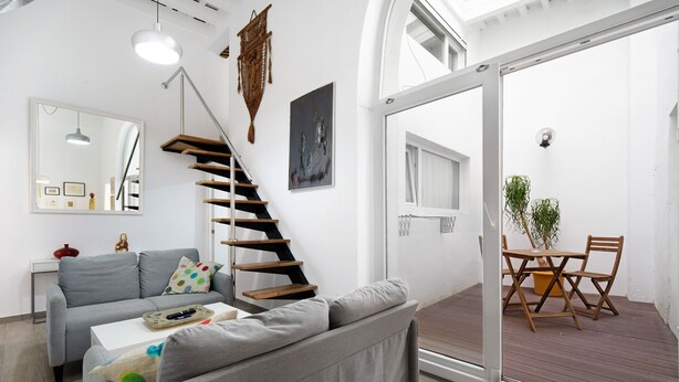 A modern living room with a grey sofa, a white coffee table, a wooden staircase, and a dining area with a table and chairs.