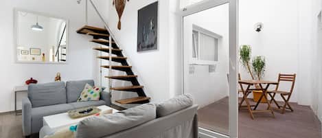 Een moderne woonkamer met een grijze bank, een witte salontafel, een houten trap en een eethoek met een tafel en stoelen.