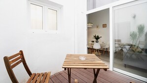 A wooden deck with a table and a chair, a white wall, and a sliding glass door leading to an interior space.