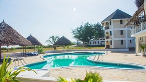 Una piscina con solárium de piedra, rodeada de sombrillas de paja y un edificio con balcones.