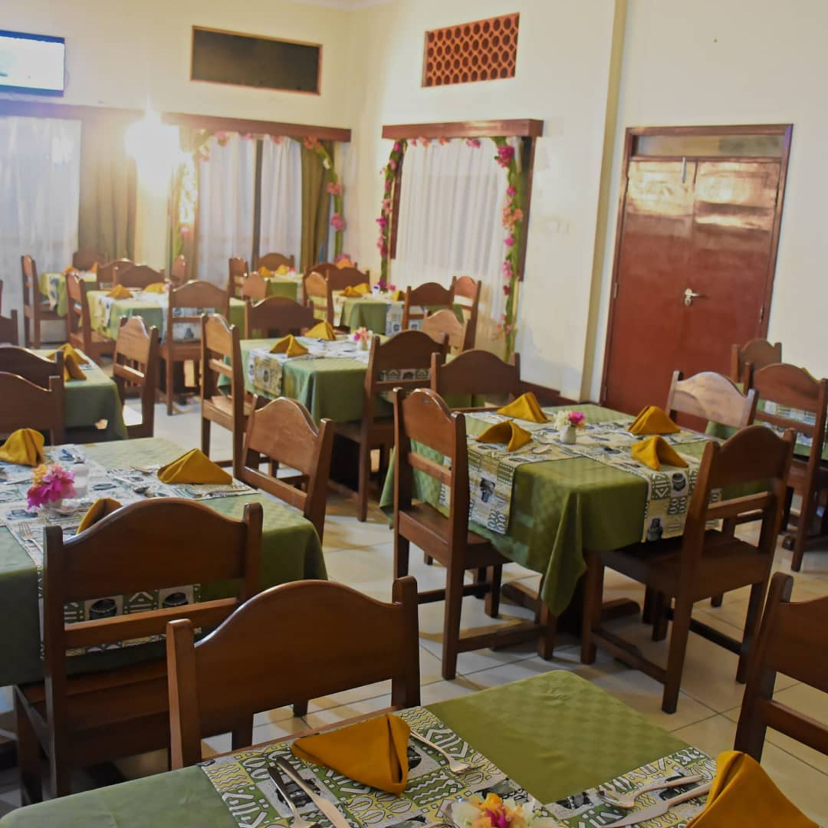A dining area with multiple tables, each covered with a green tablecloth and set with yellow napkins. The room has wooden chairs and a tiled floor.