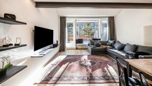 Ein modernes Wohnzimmer mit Flachbildfernseher, einer Couch, einem Esstisch und einem großen Fenster mit Blick auf Bäume.