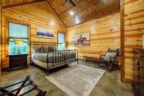 Ein gemütliches Blockhaus-Schlafzimmer mit einem Metallbettgestell, einem Schaukelstuhl, einer Holzkommode und einem großen Fenster mit Jalousien.