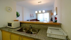 Una cocina con microondas, lavamanos y frutero. Se observa una zona de comedor con un sofá y una mesa al fondo.