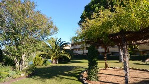 Un jardín bien cuidado con varios árboles y arbustos, un cielo despejado y un edificio con múltiples balcones al fondo.
