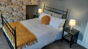 A neatly made bed with a white duvet, a brown throw blanket, and a single brown pillow. There are two bedside tables with lamps, a black metal headboard, and a floral wallpaper pattern. - Family friendly, 3 bedrooms with parking & garden. Close to beach and town. (Bridlington)