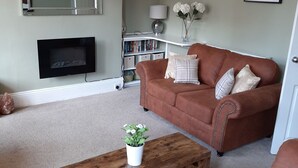 A living room with a brown sofa, a wooden coffee table with a plant, a bookshelf, and a flat-screen TV. - Family friendly, 3 bedrooms with parking & garden. Close to beach and town. (Bridlington)