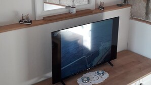 Ett vardagsrum med en platt-TV placerad på ett träskåp, en vägg av sten, en klocka och en hylla med böcker och blommor.