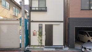 A modern two-story building with a flat facade, a small entrance with a door, and a car parked in the adjacent driveway.