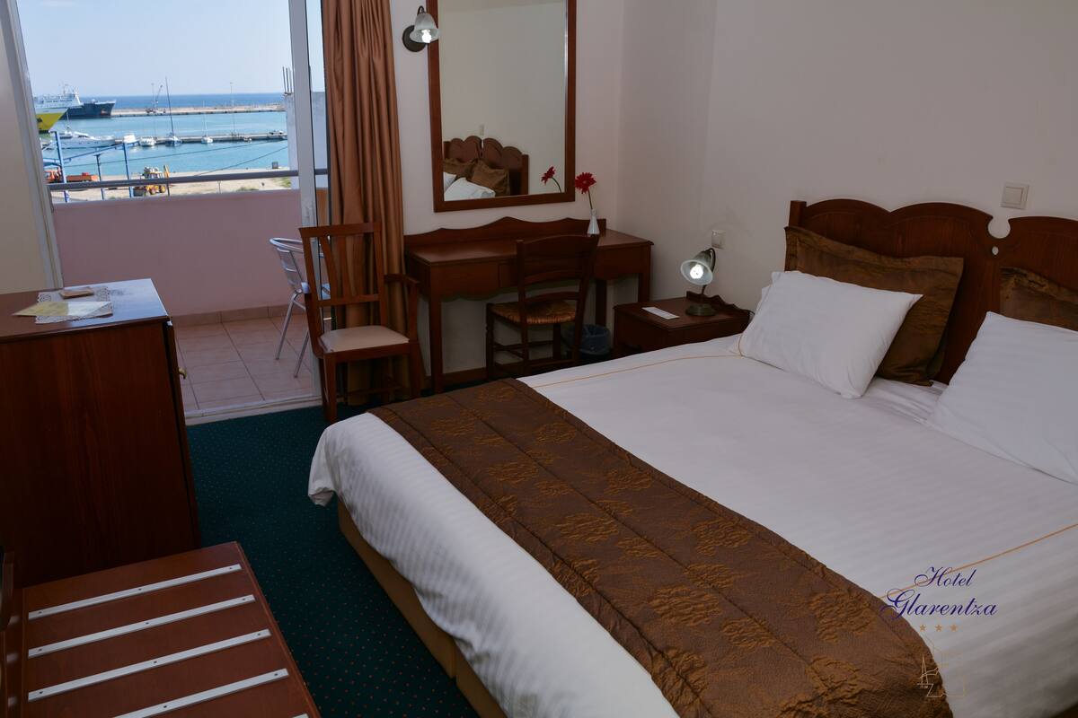 A hotel room with a large bed, a desk, a chair, a dresser, and a view of the sea.