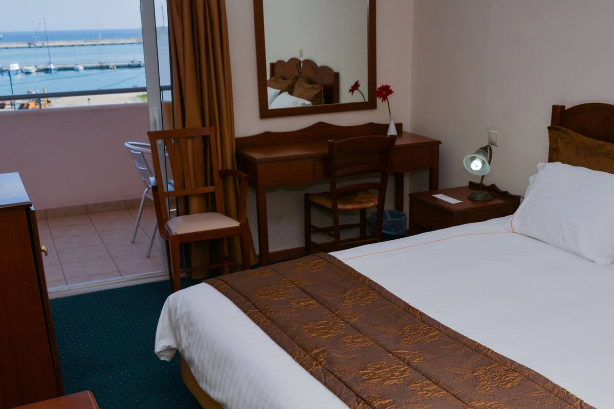 A hotel room with a bed, a desk, a chair, a nightstand, a mirror, and a view of the sea.