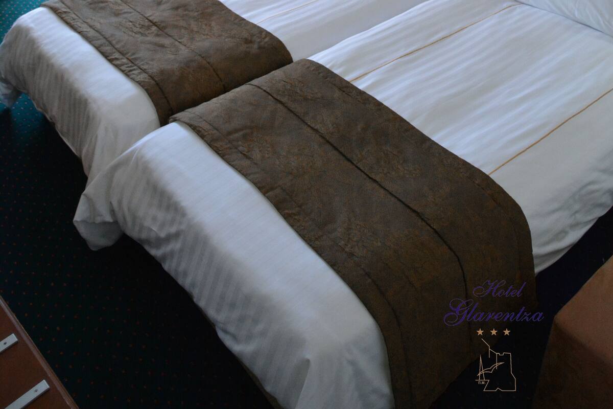 A hotel room with two beds, a chair, a small table, and a lamp.