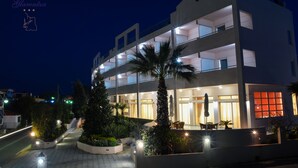 Un hotel de varios pisos con diseño moderno, que cuenta con balcones, una entrada bien iluminada y un patio ajardinado con palmeras y plantas.