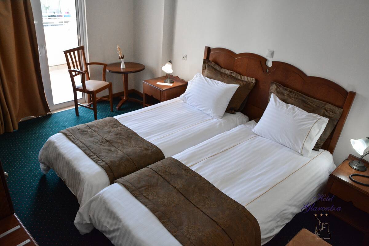A hotel room with two beds, a chair, a small table, and a lamp.