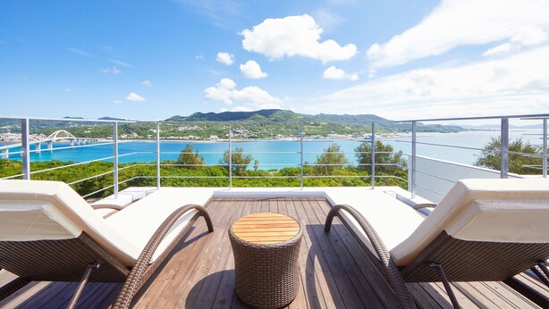 A deck with two wicker lounge chairs and a small wooden table, overlooking a coastal area with a bridge and mountains.