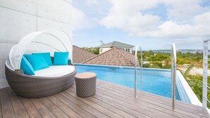 A poolside area with a wicker lounge chair, a small round table, and a view of a swimming pool and surrounding landscape.