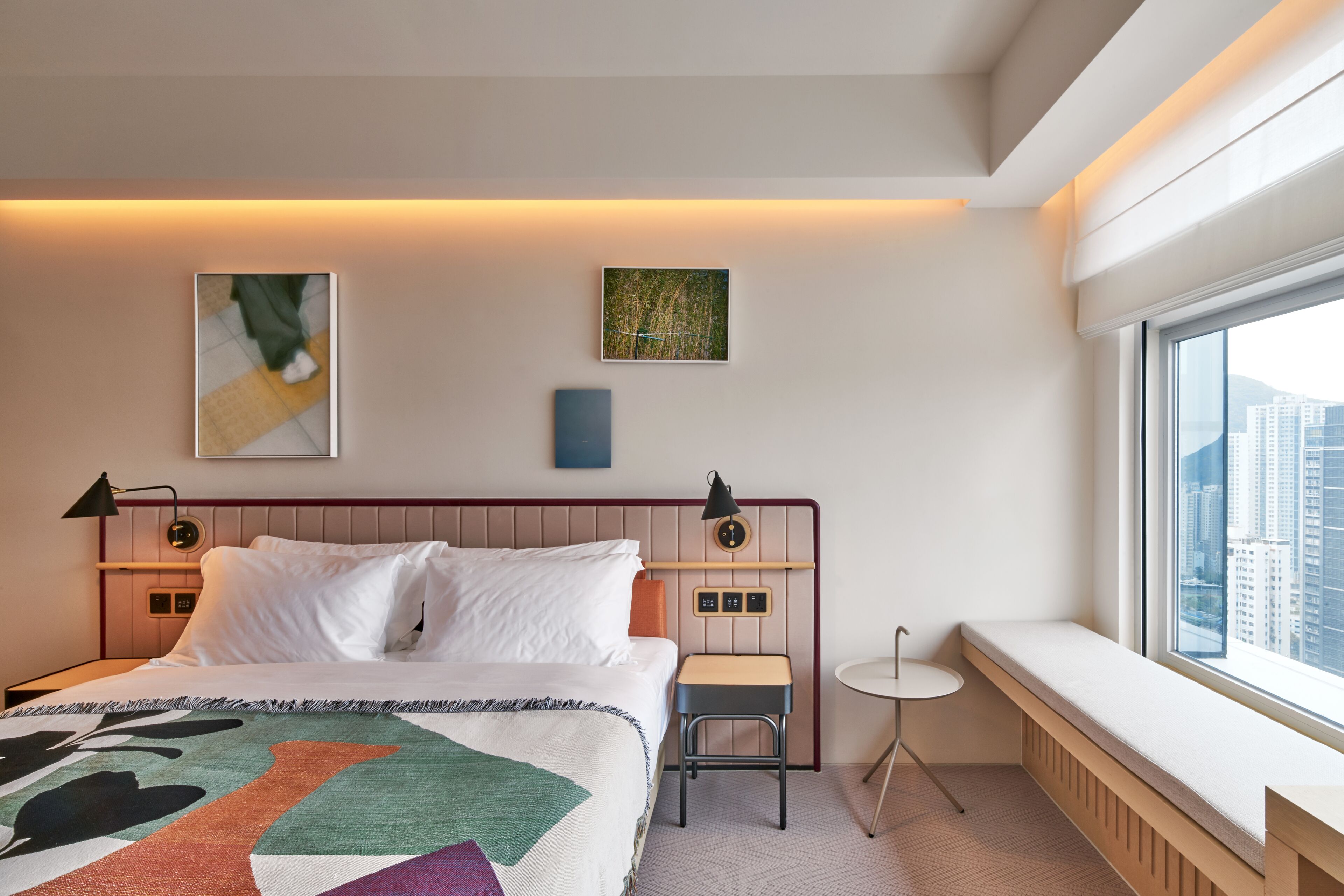 Une chambre d’hôtel moderne avec un grand lit, une petite table, un banc près de la fenêtre et des œuvres d’art accrochées aux murs.