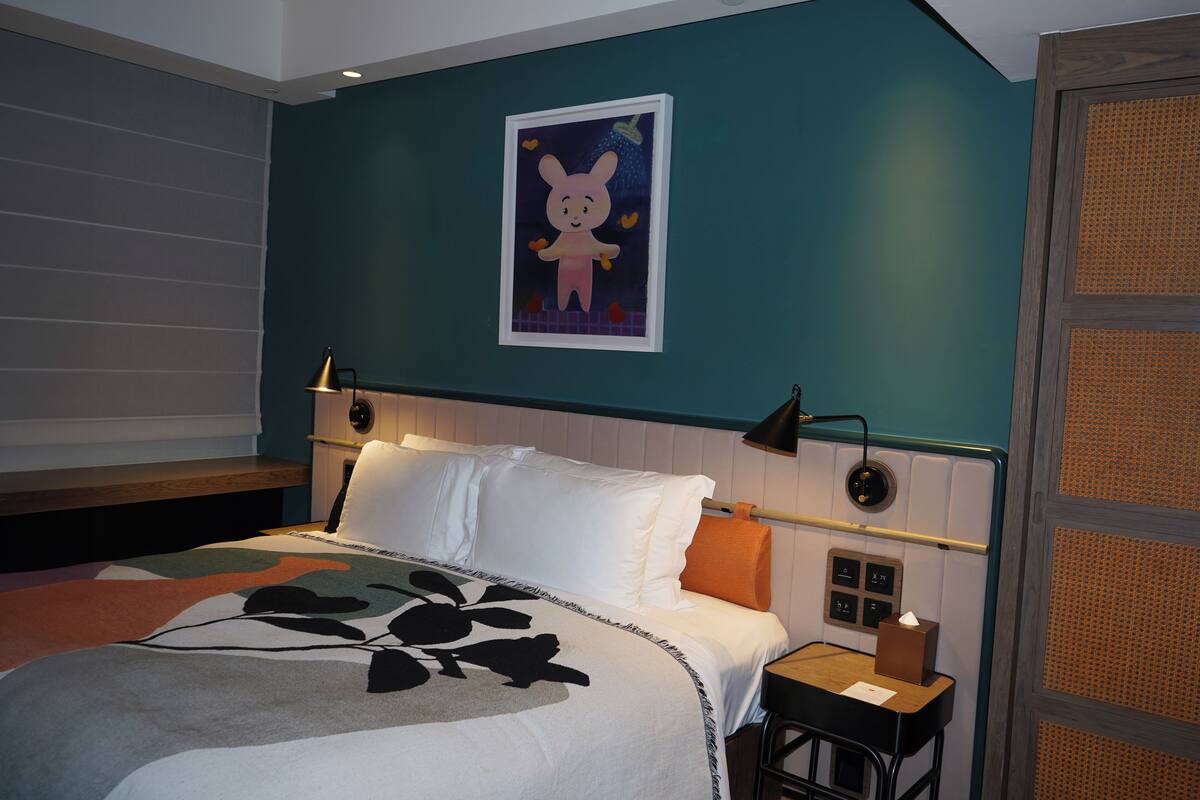 Una cama bien tendida con una colcha estampada con motivos florales, un cuadro de un personaje de caricatura en la pared y una mesita de noche con una lámpara.