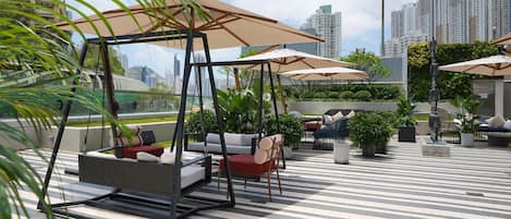 A rooftop patio with outdoor seating, including sofas and chairs, under large umbrellas, with a city skyline in the background.