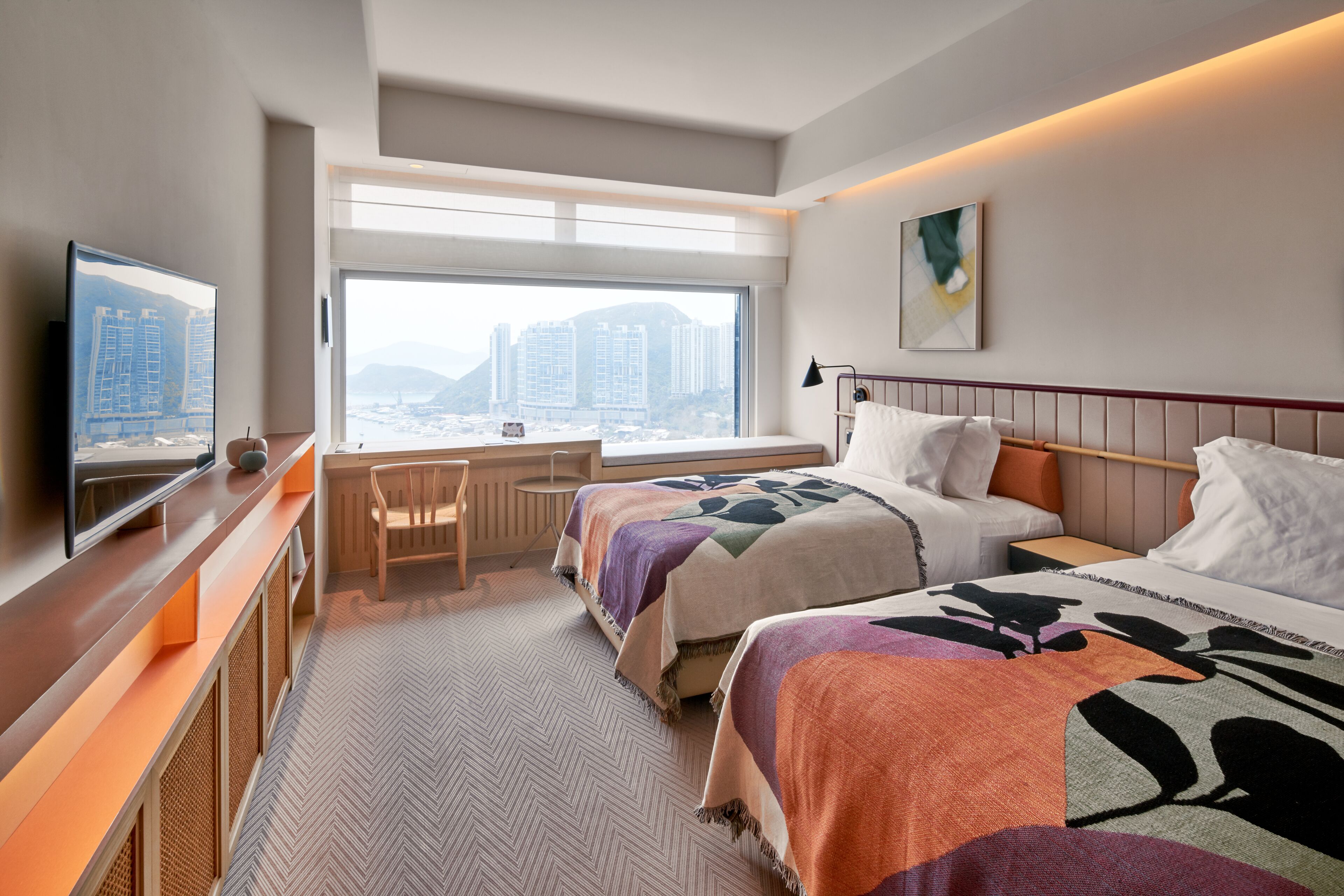 A hotel room with two beds, a desk, a TV, and a large window with a city view.