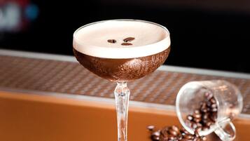 A glass with a frothy top, a coffee bean, and a spilled coffee container on a brown surface.